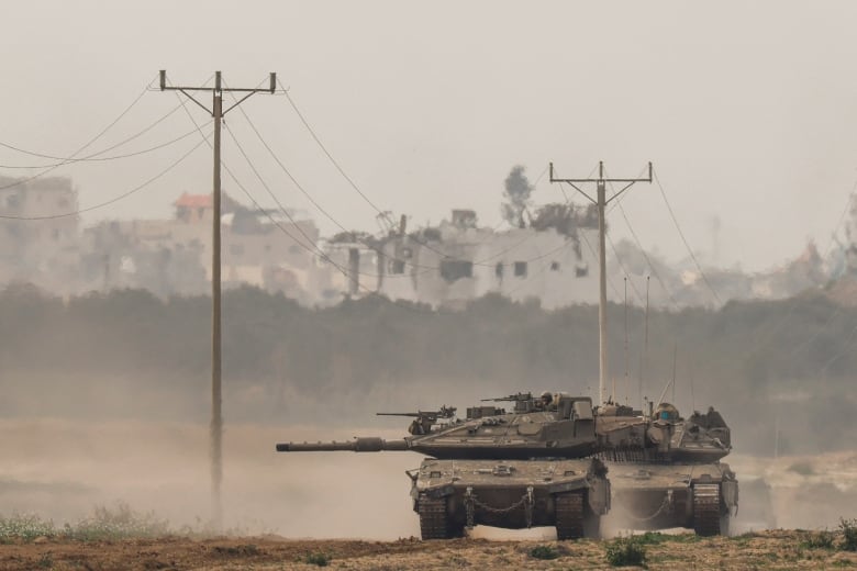 A beige tank drives on dirt, with buildings and power lines in the background.