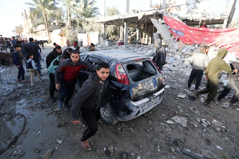 People walk next to a damaged vehicle, near rubble.